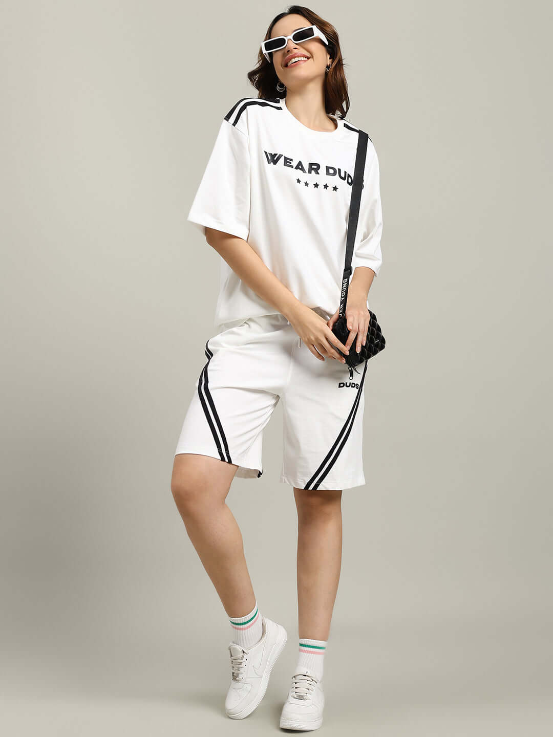 Woman wearing an oversized white t-shirt and shorts, styled with sunglasses and a bag, featuring sporty black stripes.