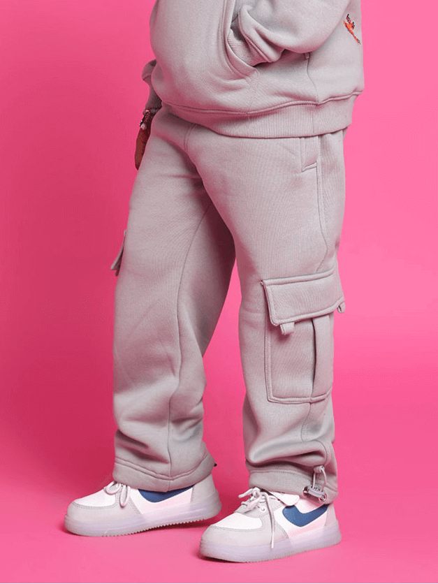 Boy wearing grey fleece pants with cargo pockets, standing against a pink background, styled casually with white sneakers.