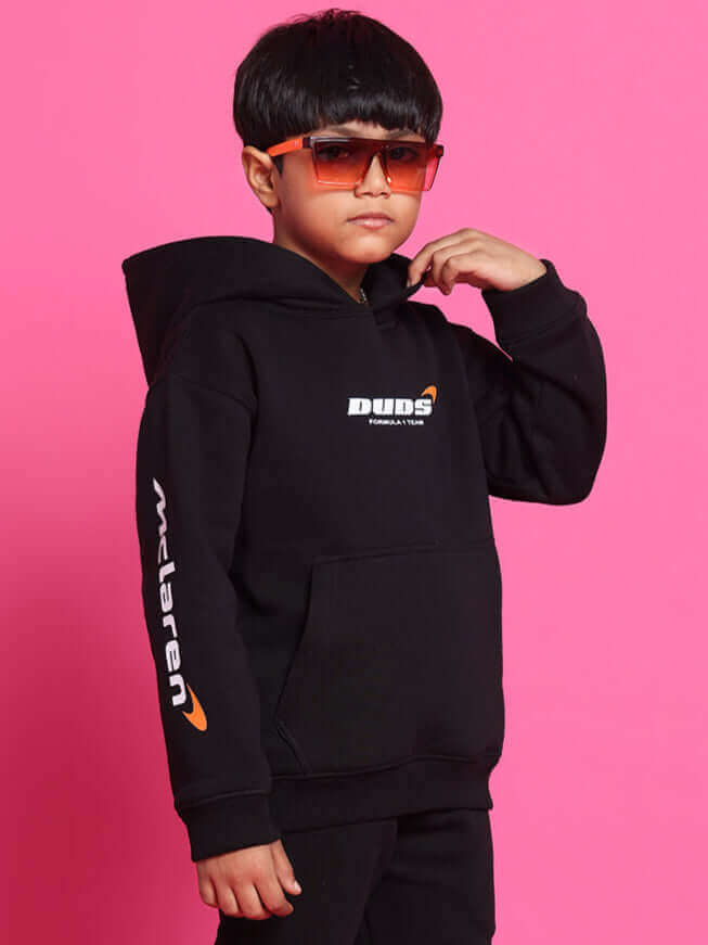 Child wearing a black monster co-ord outfit with orange sunglasses, featuring brand logos on a bright pink background.