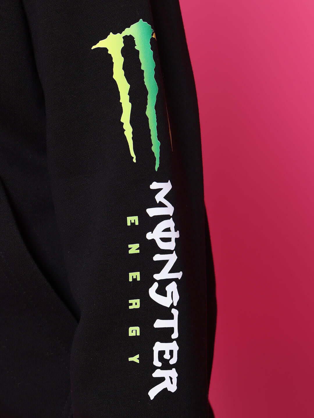 Close-up of Monster Energy logo on a black hoodie sleeve against a pink background.