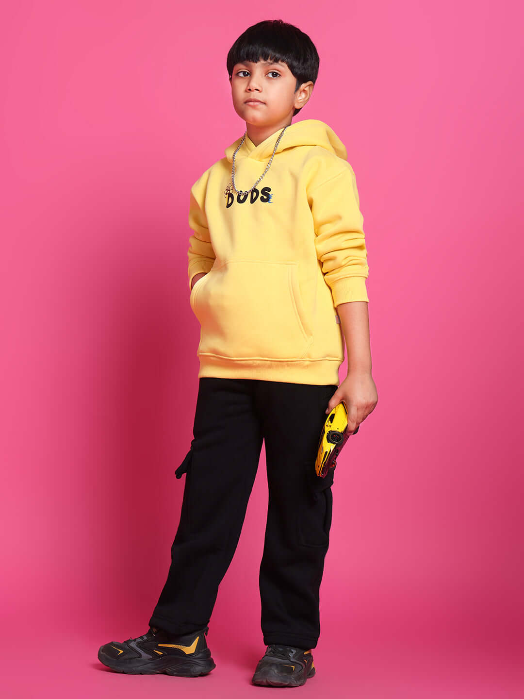 Child wearing COOL DUDE yellow co-ord outfit with hoodie and pants, posing with a toy, against a pink background.