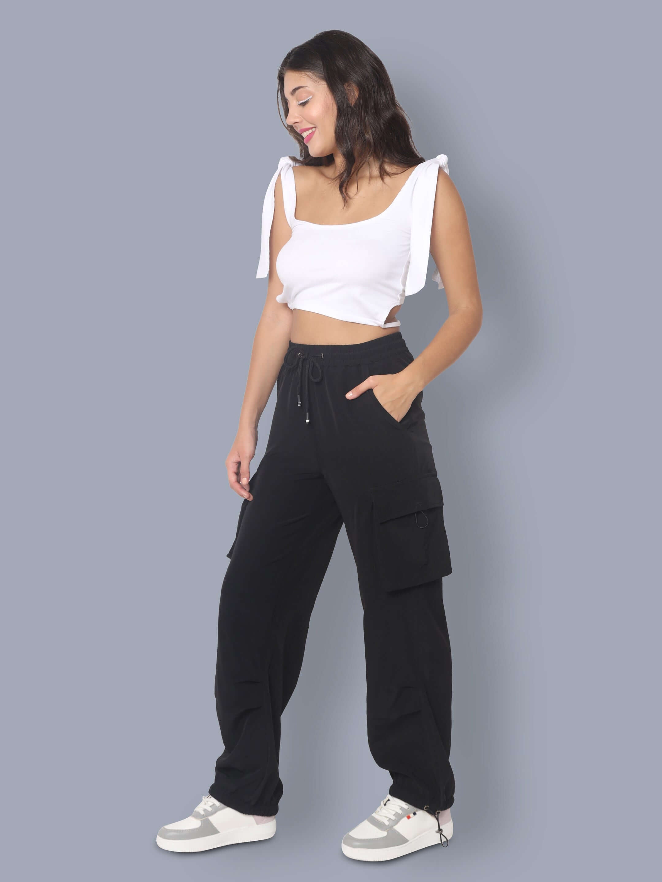 Woman wearing black tactical cargo joggers with pockets, paired with a white crop top and sneakers, standing against a gray background.
