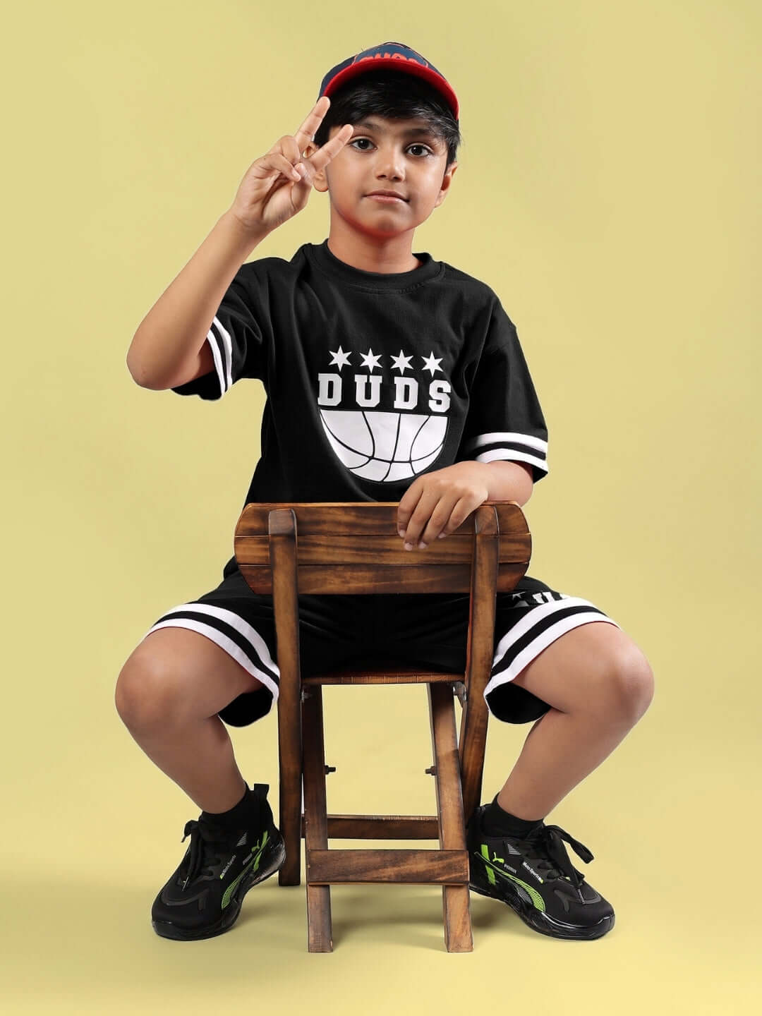 Young boy wearing a black DIGGIN oversized t-shirt from DUDS sitting on a chair, making a peace sign on a yellow background.