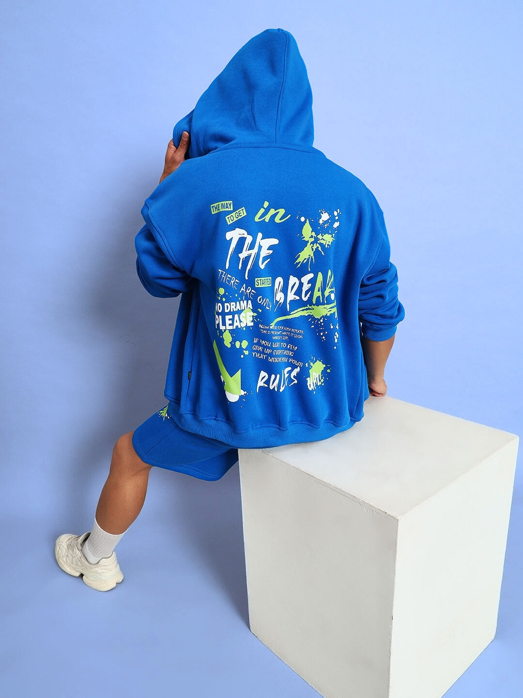 Person wearing a NO DRAMA FLEECE CO-ORD in royal blue with graphic design on the back, sitting on a white cube against a blue background.