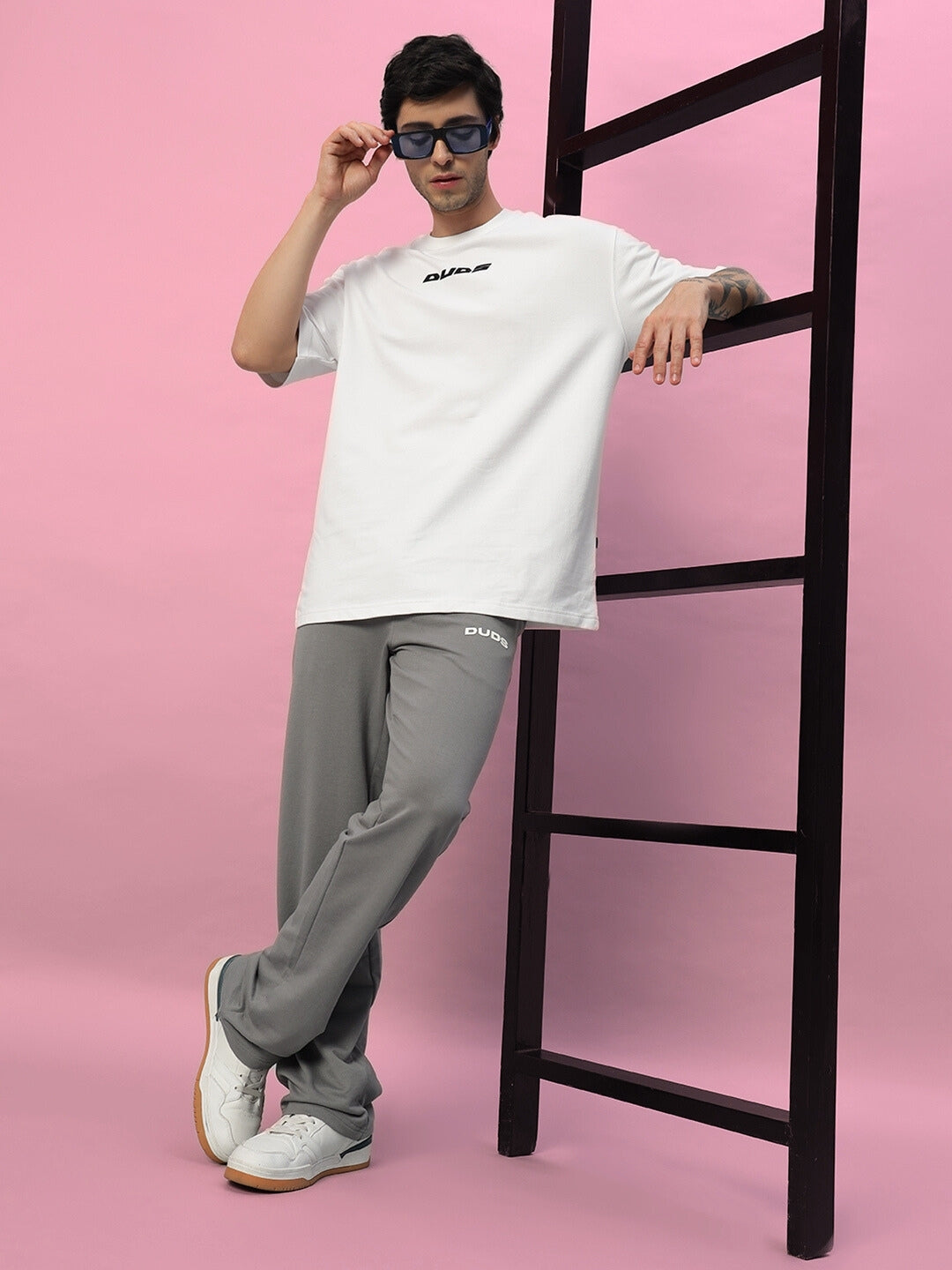 Model wearing New Era Relaxed Fit Grey Joggers by DUDS, styled with a white t-shirt and sunglasses, posing against a pink background.