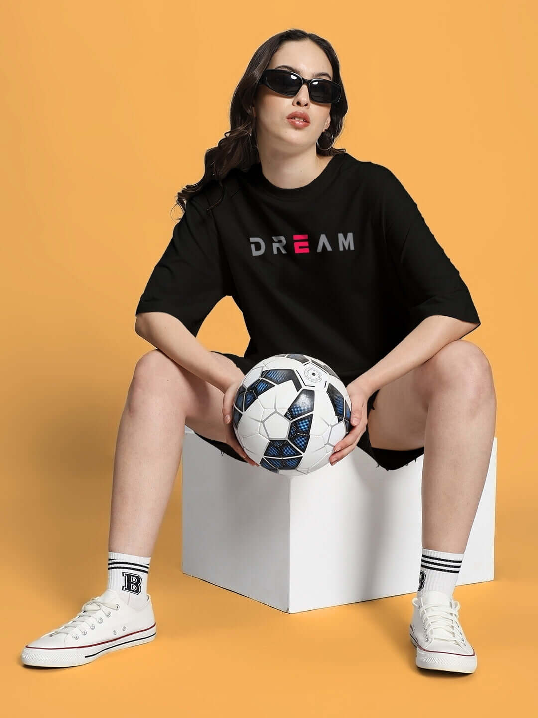 Woman in black shirt and shorts holding soccer ball sitting on box against orange background.