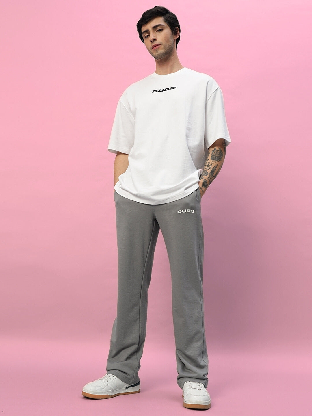 Person wearing New Era Relaxed Fit Grey Joggers by DUDS, standing against a pink background. Casual and stylish made-to-order apparel.