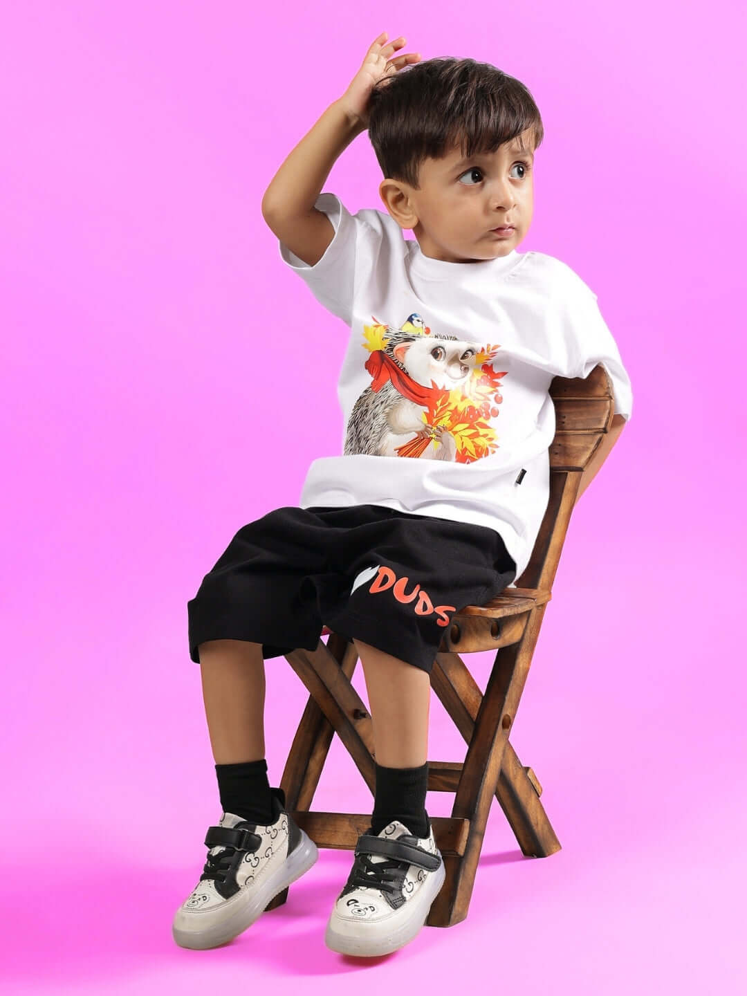 Child wearing a white hedgehog-themed T-shirt by DUDS, sitting on a wooden chair against a pink background.