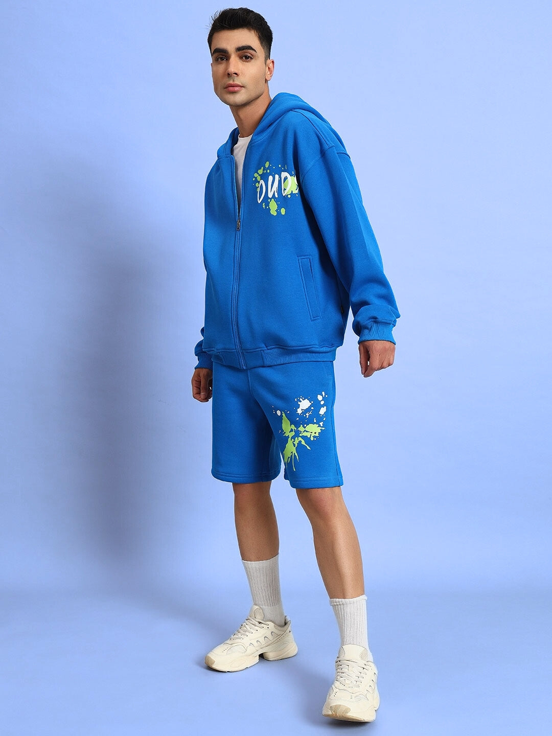 Model wearing NO DRAMA fleece co-ord in royal blue with graphic print, featuring a hoodie and shorts, paired with sneakers.