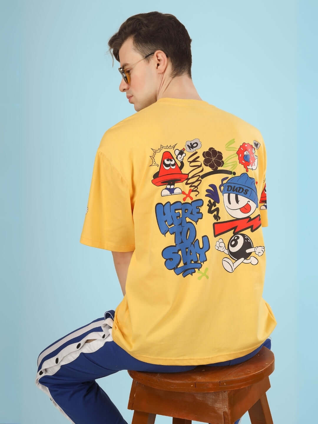 Man wearing HERE TO STAY OVER-SIZED T-SHIRT YELLOW with graphic design, sitting on stool, showcasing bold street style fashion.