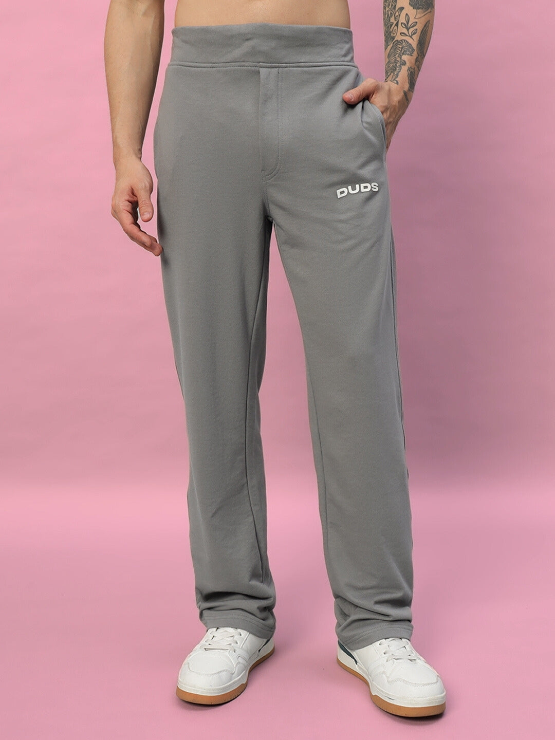 Man wearing grey DUDS relaxed fit joggers with white sneakers against a pink background.
