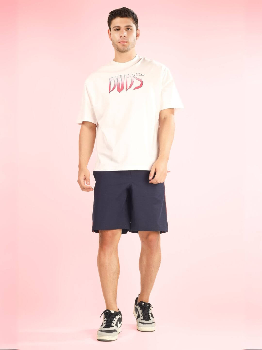 Model wearing CRUEL OVER-SIZED T-SHIRT BABY PINK by DUDS with navy shorts and sneakers, front view, pink background.