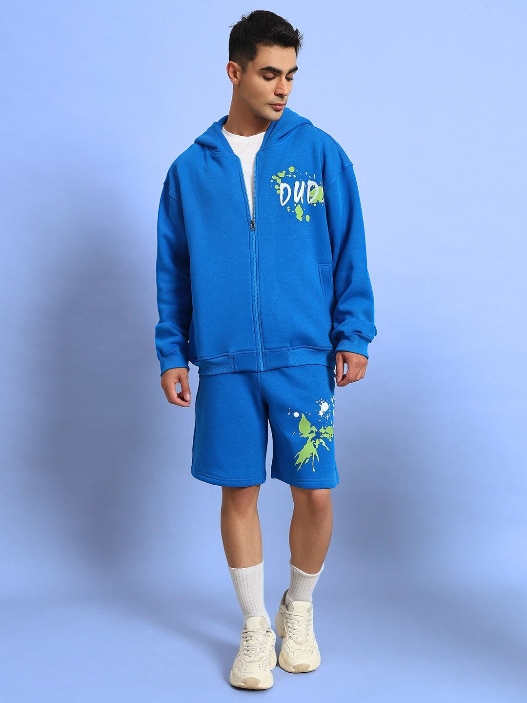 Man wearing NO DRAMA fleece co-ord set in royal blue, featuring a hoodie and shorts with graphic details, on a blue background.
