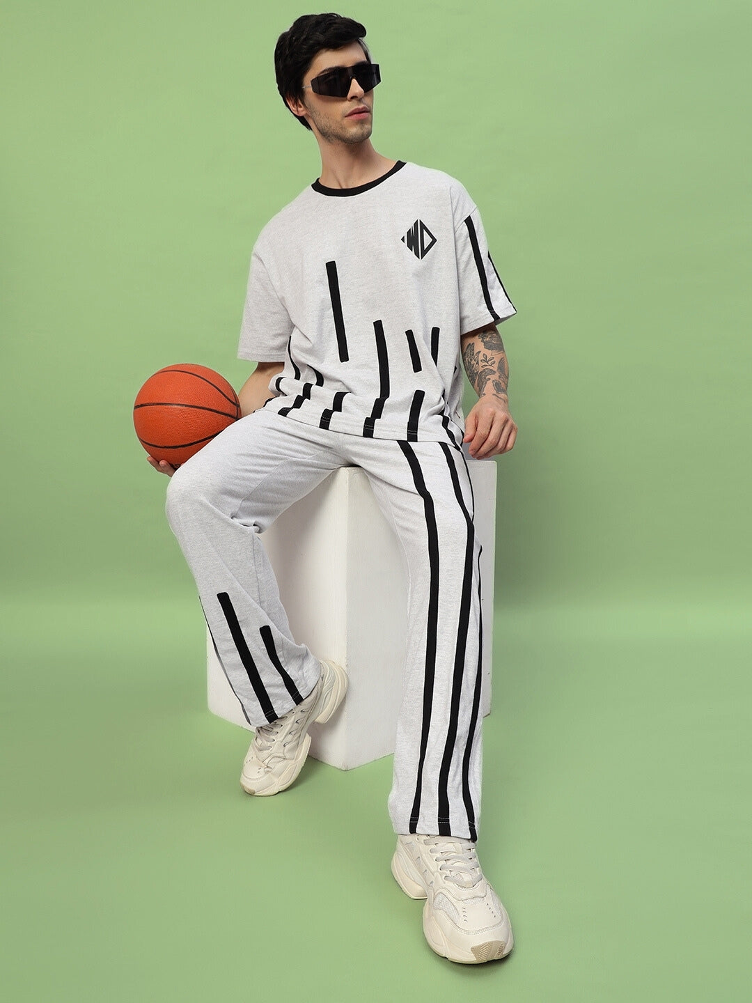 Model wearing a Bastille Oversized T-Shirt in melange grey, seated with a basketball, featuring black stripe accents and sunglasses.