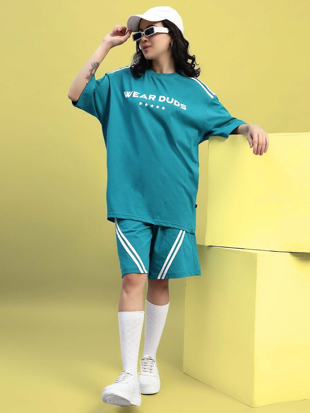Model wearing teal oversized women's star jumper T-shirt, styled with matching shorts, white cap, and sunglasses, against yellow background.
