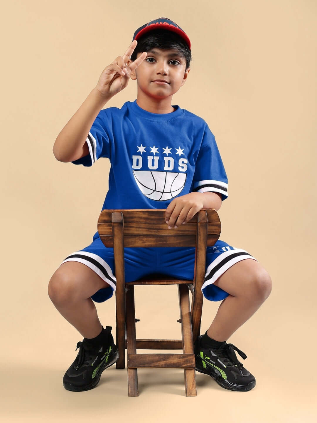Young boy posing in blue Digs shorts and shirt by DUDS, sitting on chair with peace sign, showcasing kids' athletic casual wear.