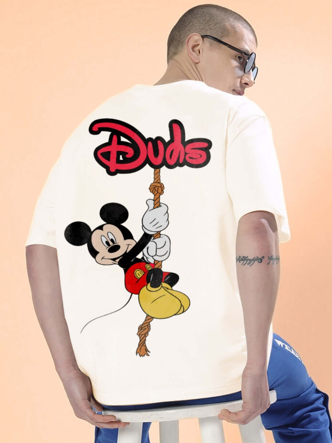 Person wearing DUDS MOUSE OVER-SIZED T-SHIRT OFF WHITE with cartoon graphic on the back, sitting on a stool.