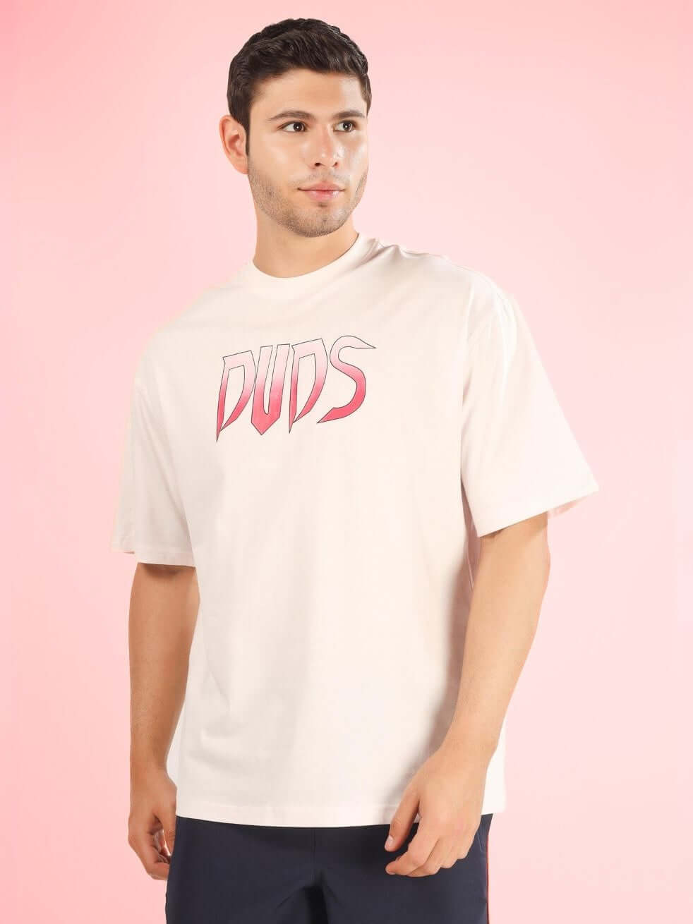 Man wearing DUDS CRUEL OVER-SIZED T-SHIRT BABY PINK with printed logo, displayed against a pink background.