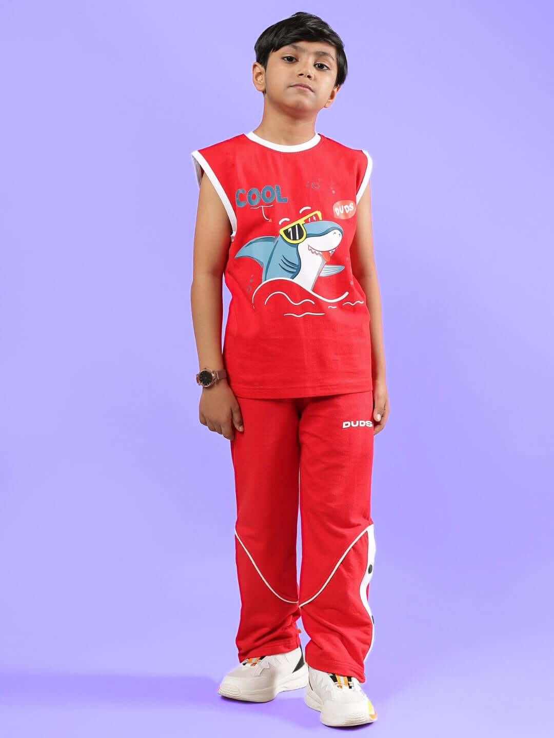 Child wearing a red unisex Shortfin t-shirt with shark graphic, paired with matching pants, from DUDS, standing against a purple background.