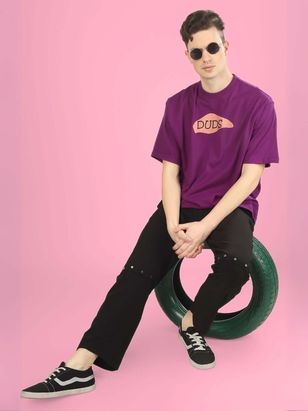 Model wearing WHAT WOW OVER-SIZED PRINTED T-SHIRT PURPLE, sitting on a tire against a pink background.