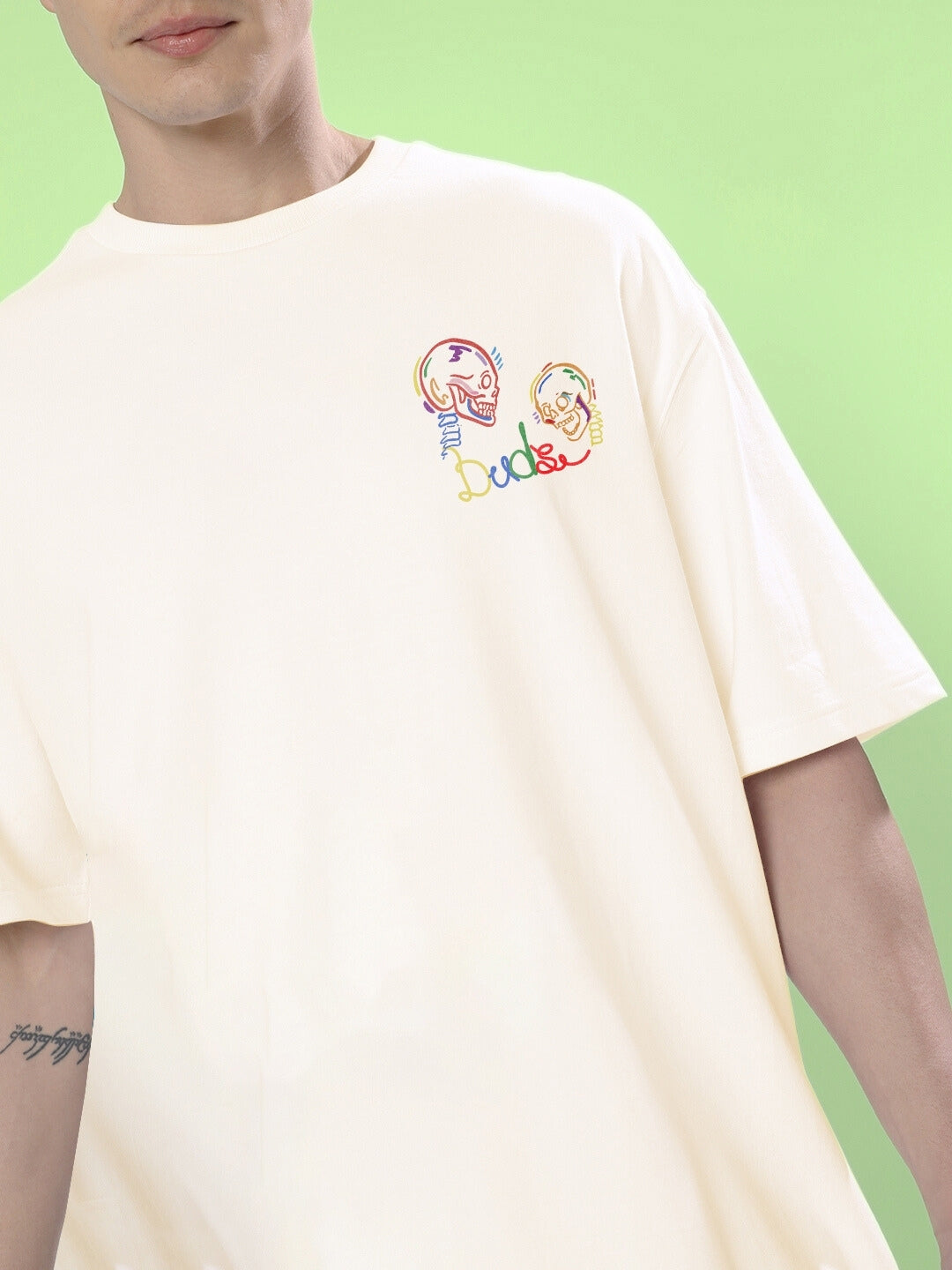 Man wearing SKULL ROLL OVERSIZED T-SHIRT OFF WHITE with colorful skull design, green background.