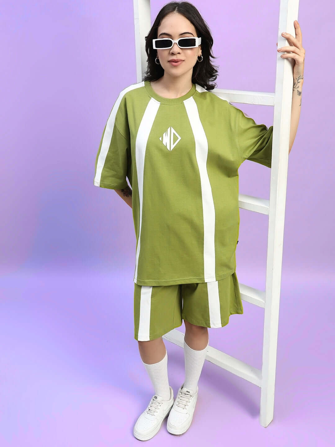 Woman wearing olive green oversized t-shirt with white stripes, styled with shorts and sneakers, standing by a ladder.