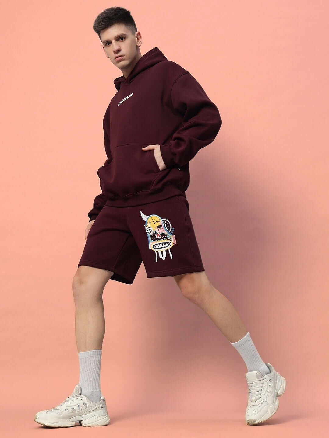 Model wearing Jumbled Fleece Co-ord in wine; stylish hoodie and shorts with graphic print, matched with white sneakers on pink background.