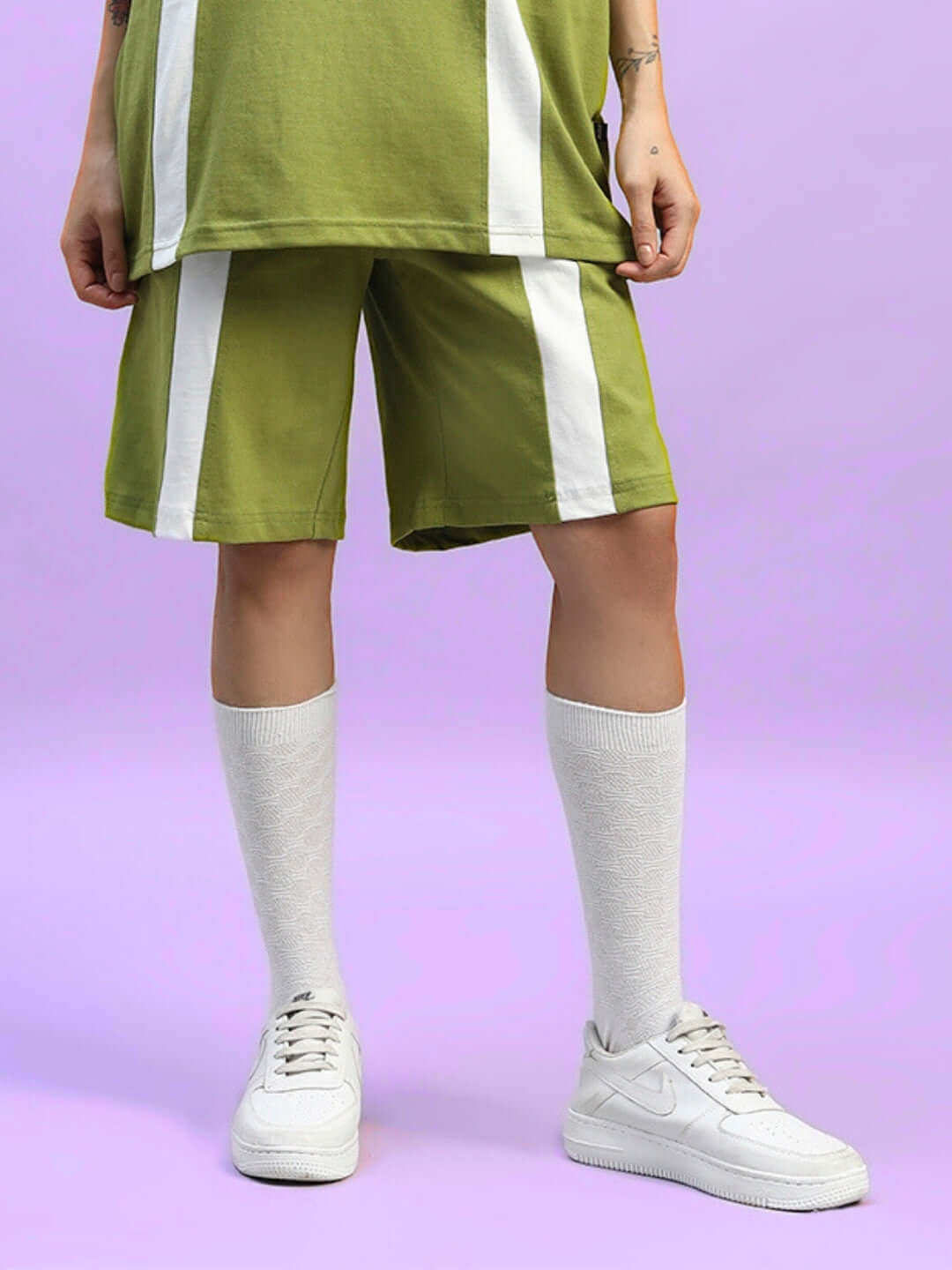 Woman wearing olive green wideline regular fit shorts with white stripes, paired with white knee socks and sneakers, on a pastel purple background.