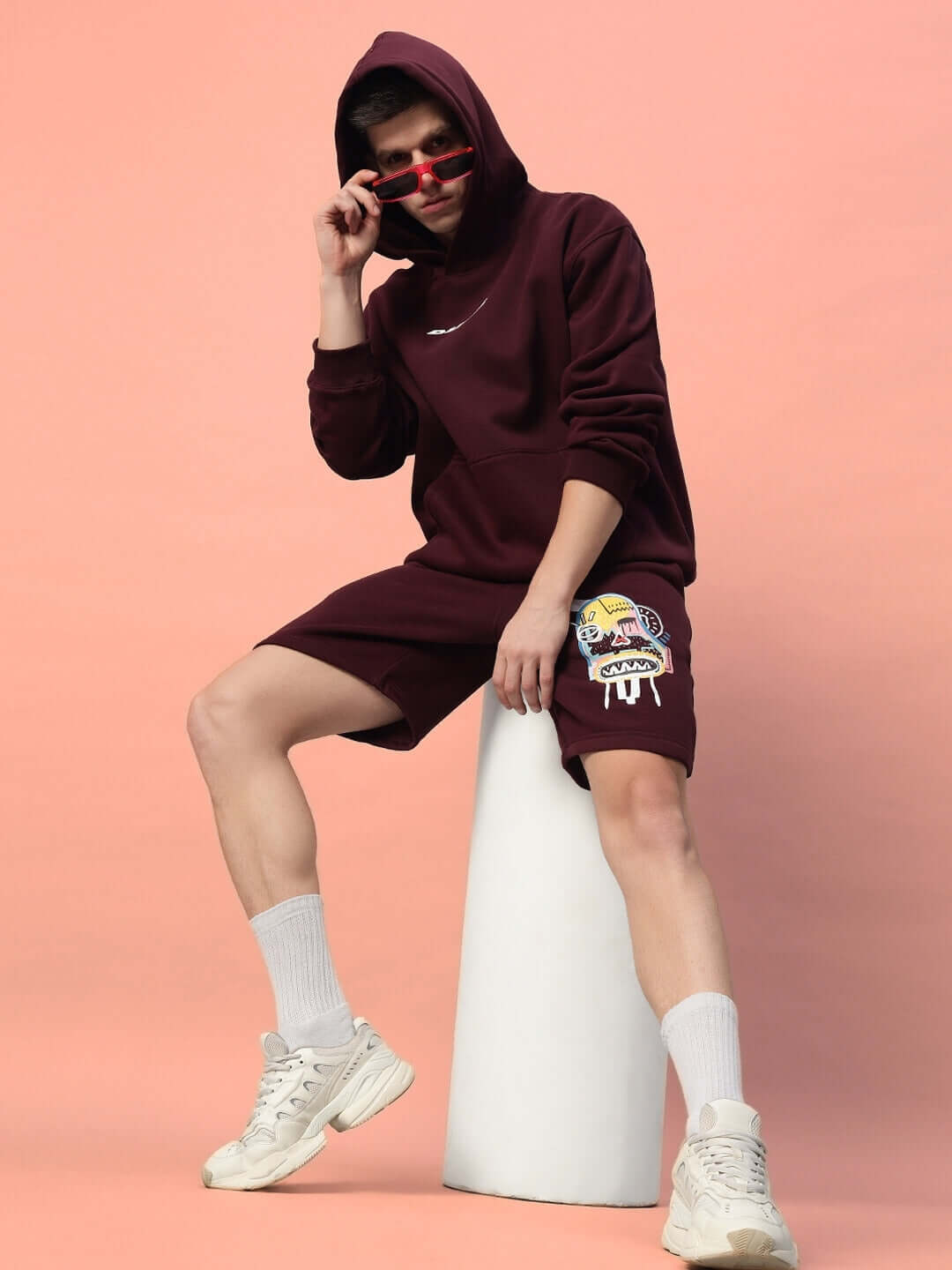 Person wearing a wine-colored jumbled fleece co-ord set with hoodie and shorts, sitting on a stool against a pink background.