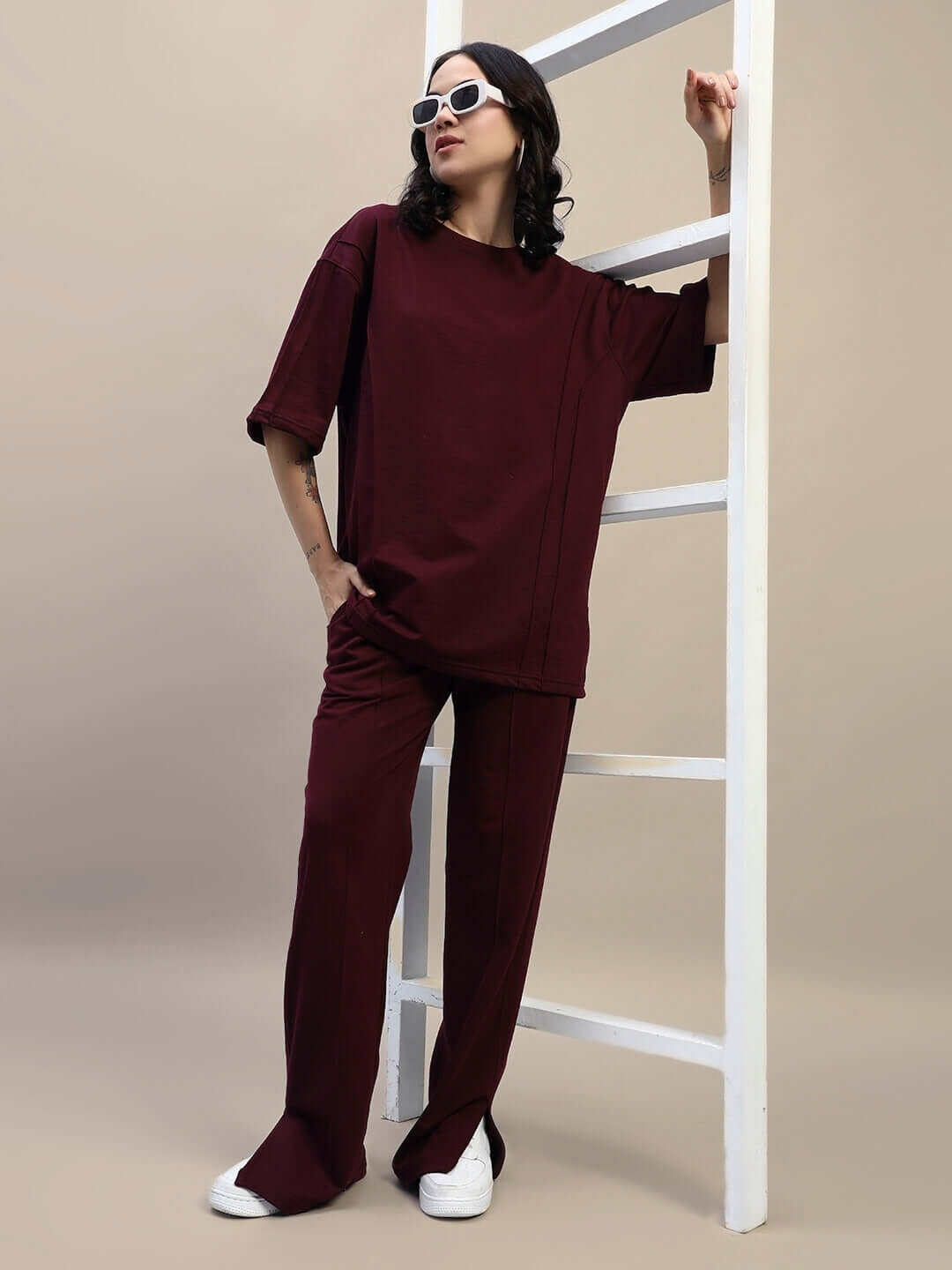 Model wearing maroon women's oversized t-shirt, premium cotton blend, styled with matching pants and white sneakers, leaning on ladder.
