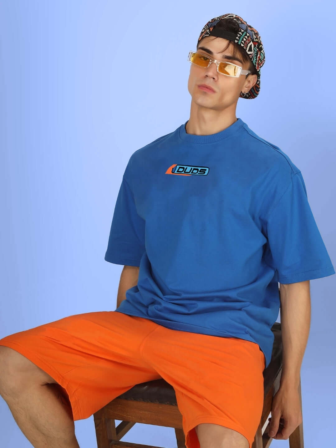 Model wearing ROBO SHARK OVER-SIZED T-SHIRT ROYAL BLUE with sunglasses, sitting on a stool.