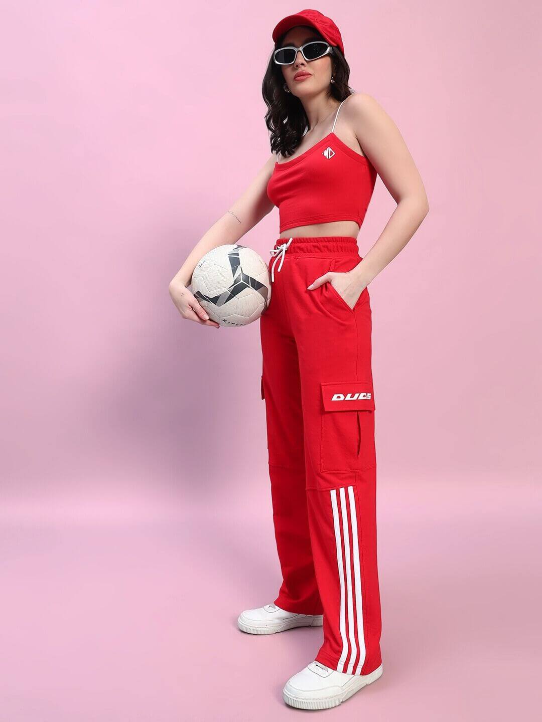 Woman in red Rosella co-ord set holding a soccer ball, featuring a crop top and cargo pants, styled with a cap and sunglasses.