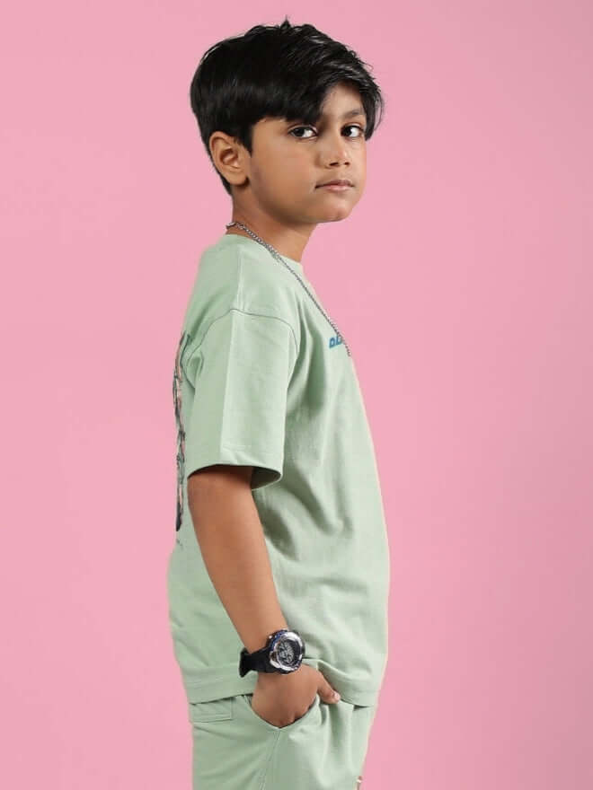 Boy wearing Nyanza Green Pray & Peace T-shirt, side view against pink background.