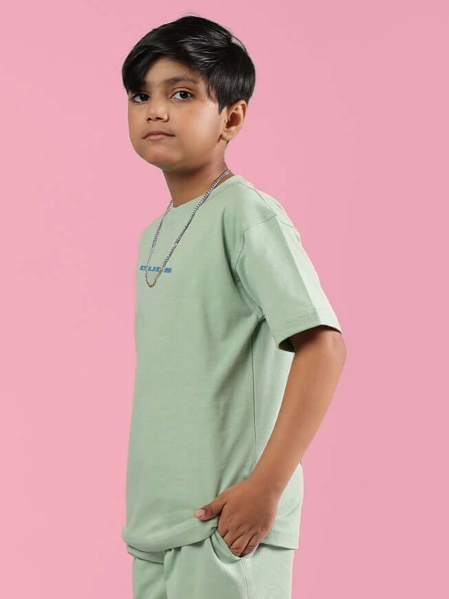 Child wearing Nyanza Green Pray & Peace T-shirt for boys and girls by DUDS, standing against a pink background.