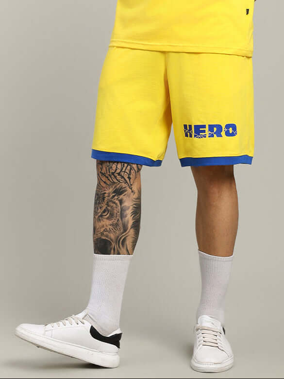 Person wearing yellow superhero-themed summer shorts with 'Hero' logo, showcasing casual style with white sneakers and lion tattoo visible.