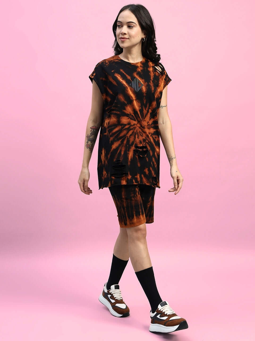 Woman wearing black ripped tie-dye co-ord set with muscle tee and shorts, styled with sneakers and socks, posing against pink backdrop.