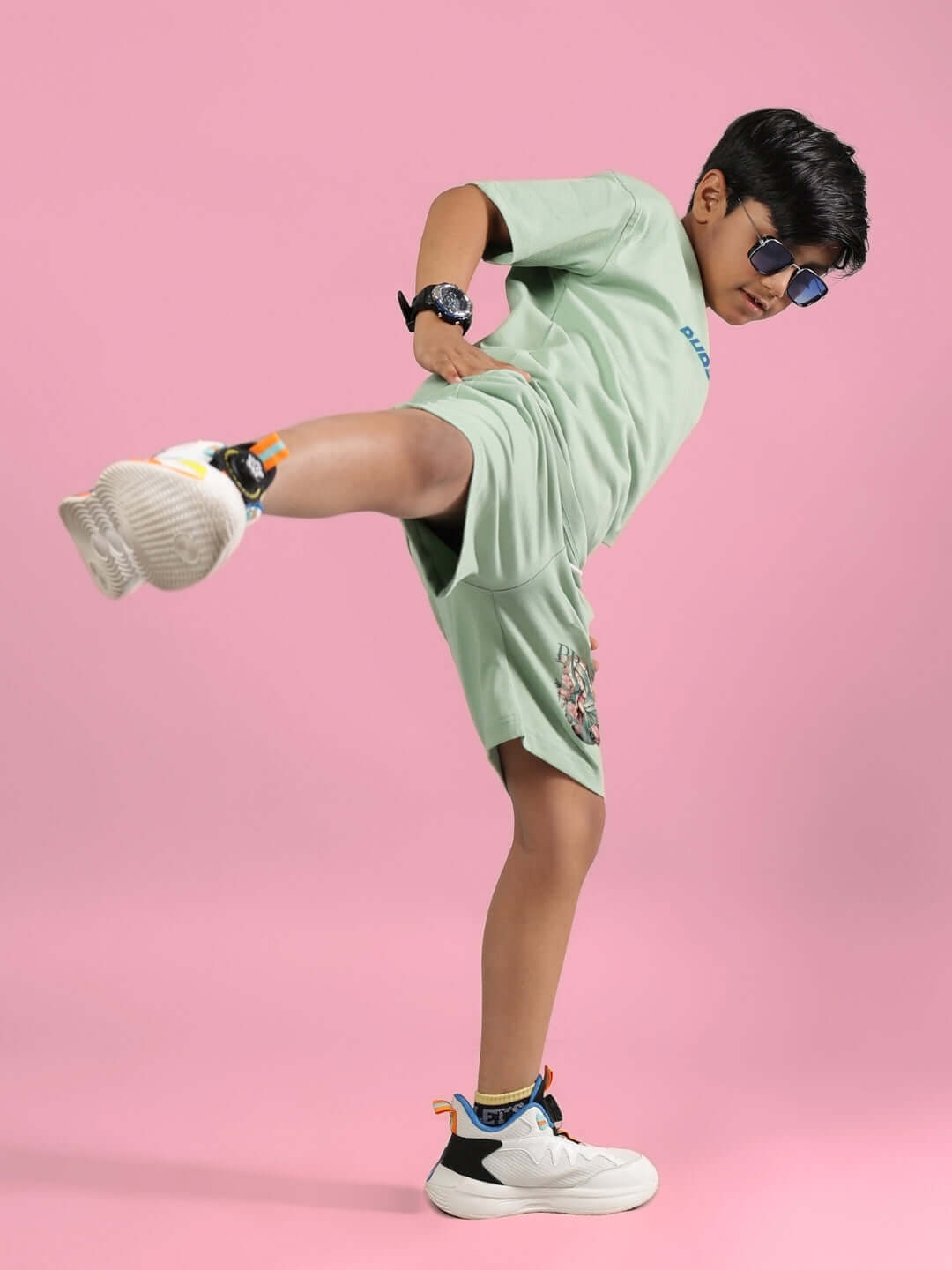 Boy wearing PRAY & PEACE NYANZA GREEN shorts performs a dynamic kick, showcasing stylish kids' clothing against a pink background.