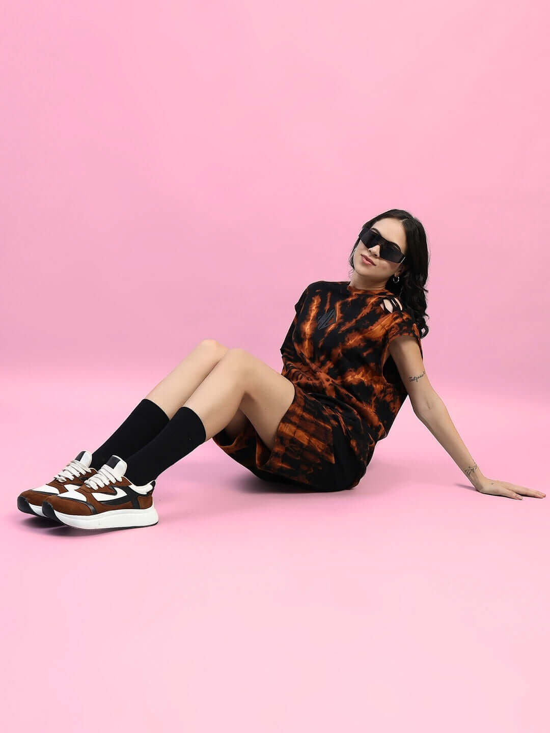 Woman wearing black tie-dye muscle tee and shorts set, lying on pink backdrop, styled with sneakers and sunglasses, showcasing casual fashion.
