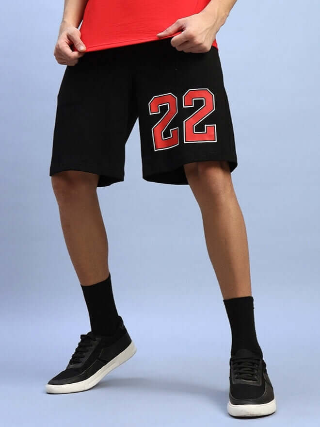 Man wearing Amiga Regular Fit Black Shorts with red '22' print, paired with black socks and sneakers, standing against a blue background.