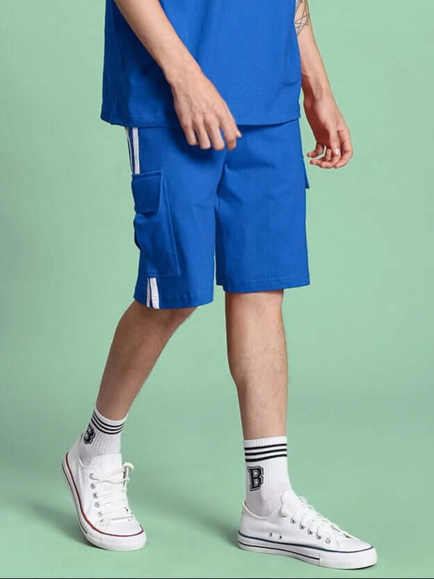 Man wearing blue Scotia regular fit shorts with side pockets, pairing them with white sneakers and striped socks against a green background.