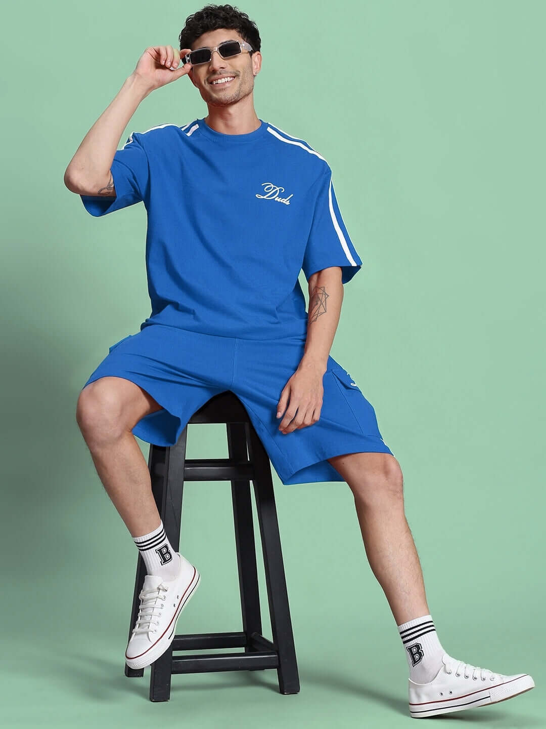 Model wearing Scotia Regular Fit Blue Shorts with matching shirt, sitting on a stool, styled with sneakers and sunglasses.