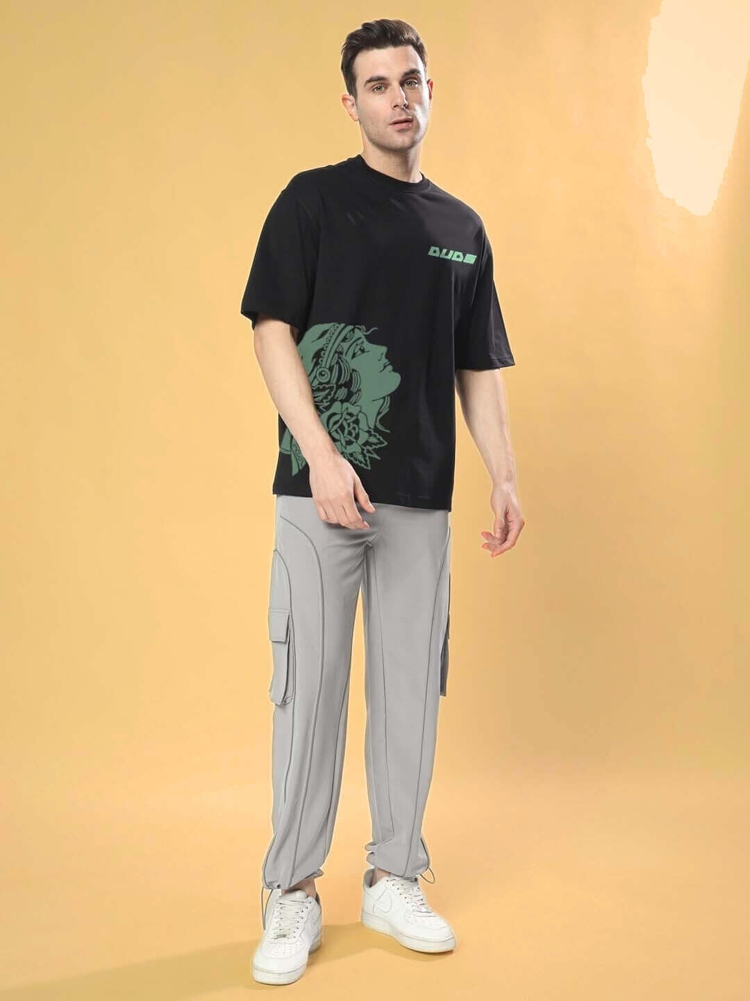 Male model wearing KINNAUR OVER-SIZED T-SHIRT BLACK with graphic print, paired with light gray cargo pants, posing against a neutral background.