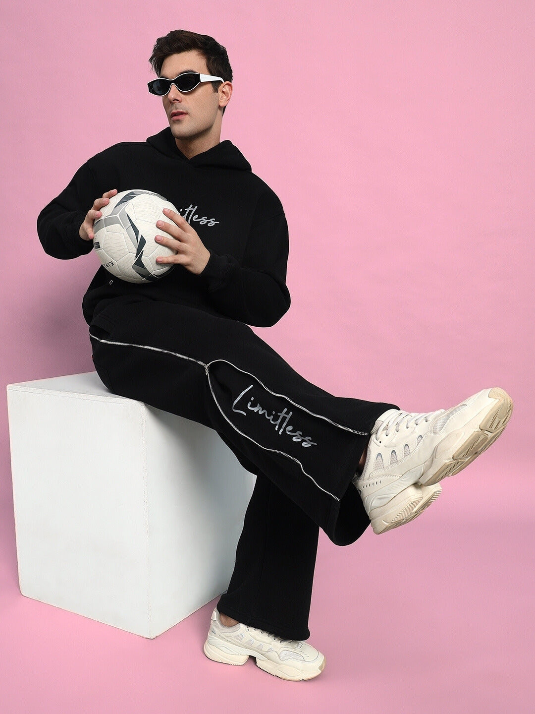 Man wearing Limitless Fleece Reflector Co-ord in black holds soccer ball, sitting against pink backdrop. Stylish and sporty look.