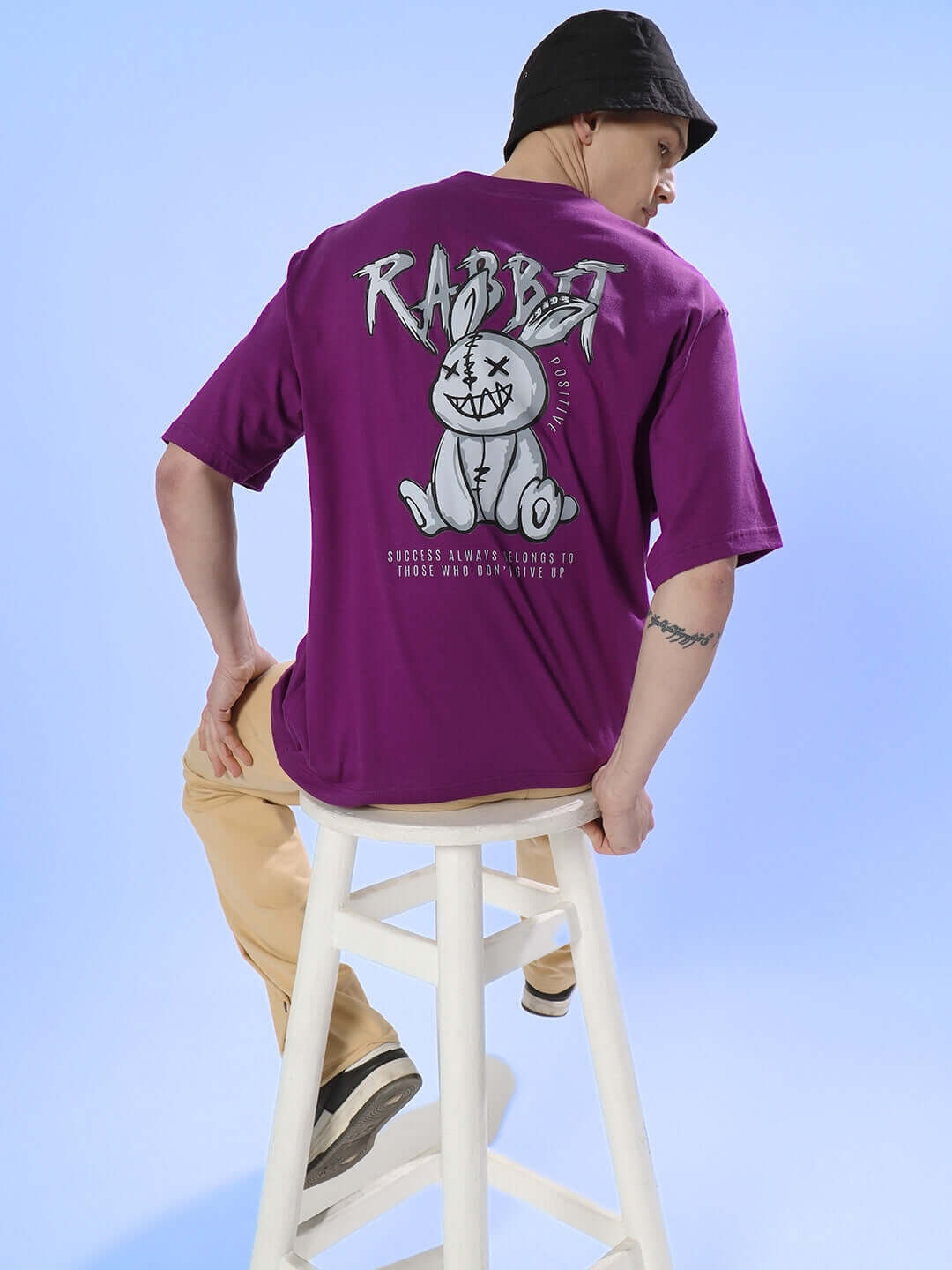 Man wearing Positive Rabbit Over-Sized T-Shirt Purple with a playful rabbit graphic, sitting on a stool against a blue background.