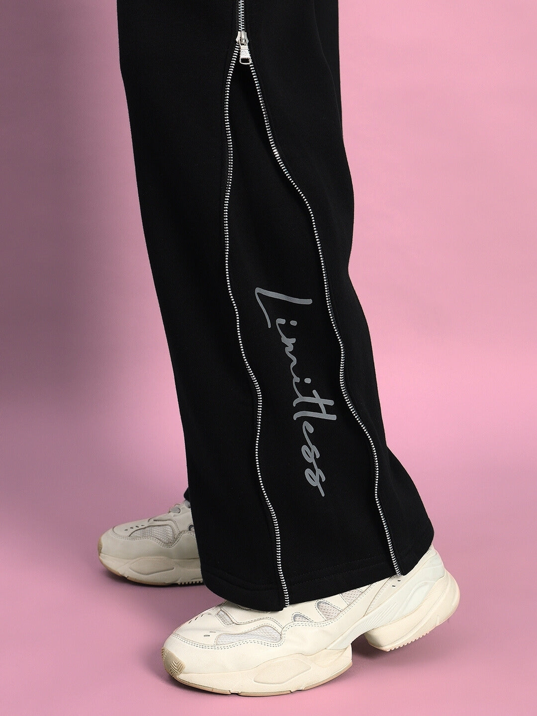 Black Limitless Fleece Reflector Jogger showcasing reflective side zippers and sporty sneakers on a pink background.
