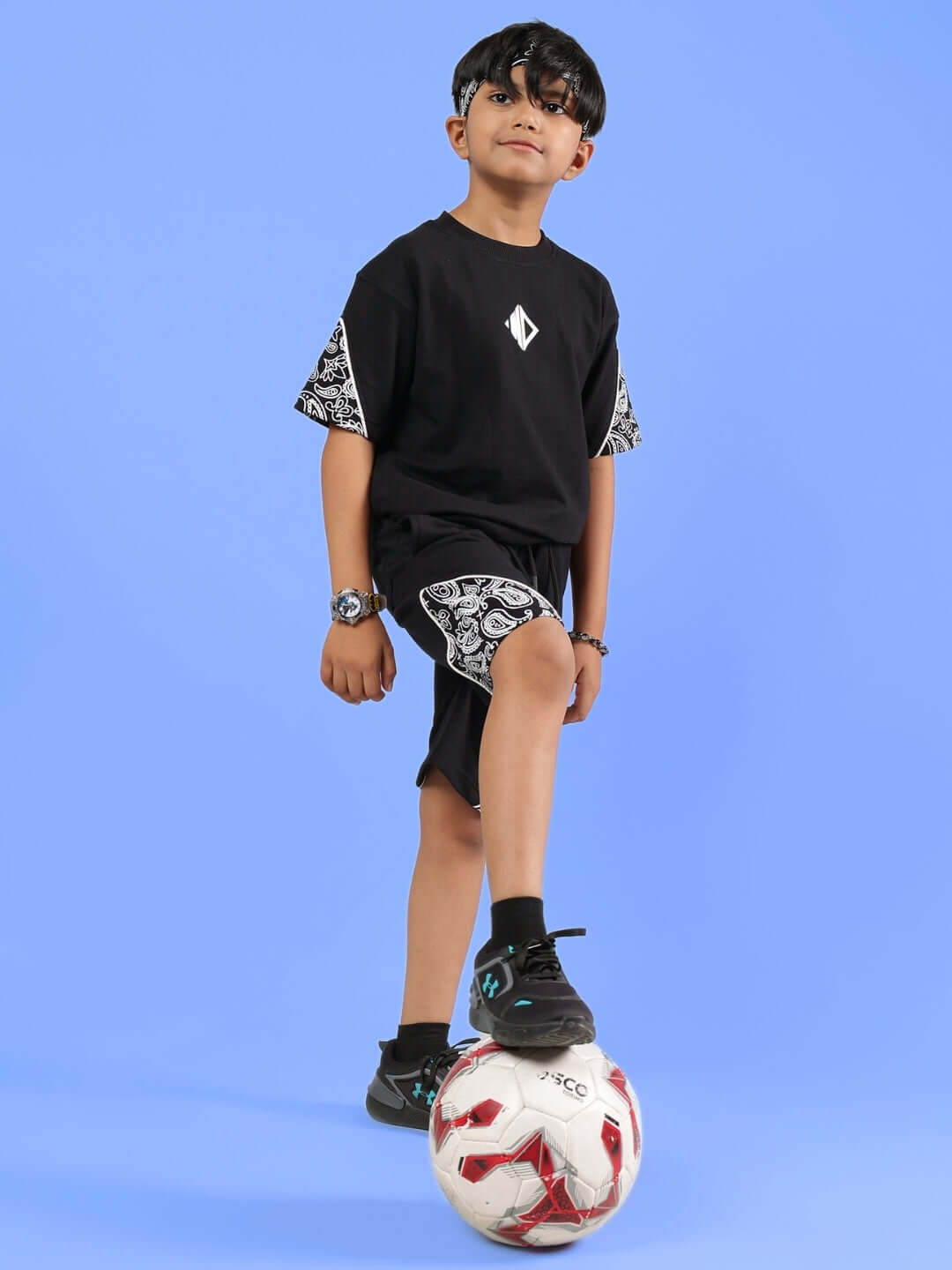 Young boy wearing a DUDS black Calvin oversized T-shirt, standing with a soccer ball on a blue background.