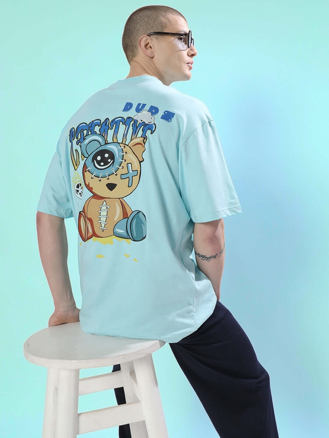 Person wearing DUDS CREATIVE OVER-SIZED T-SHIRT SKY BLUE with graphic print on back, sitting on stool.