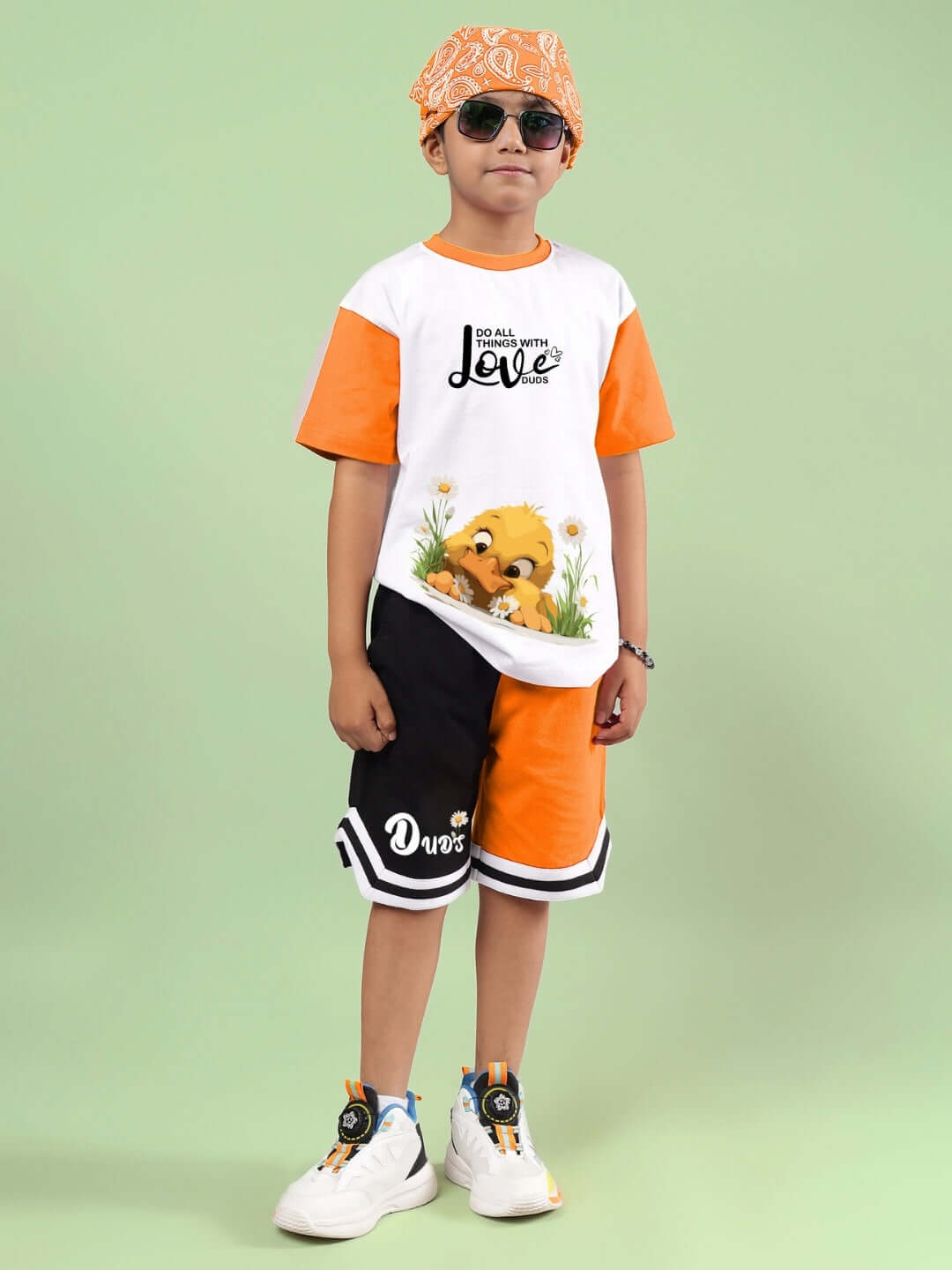 Child in white and orange Friendship Bucket Co-ord set by DUDS with a bear print, matching shorts, cap, and sneakers.