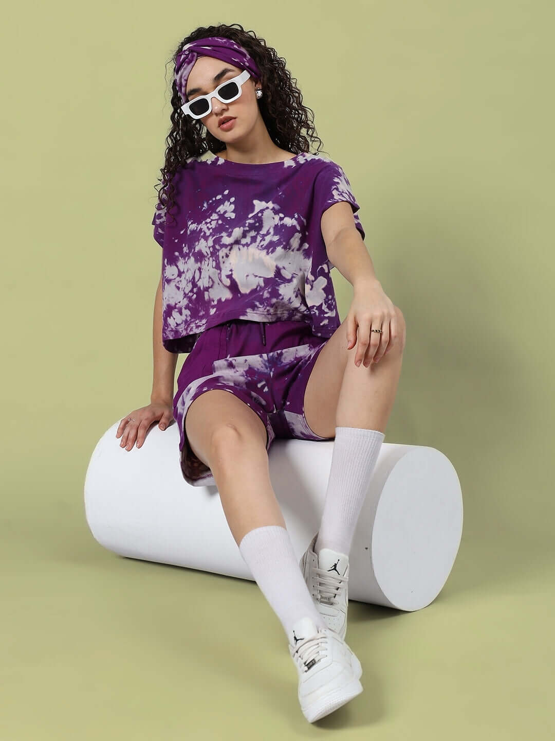 Woman in purple tie-dye co-ord set with headband and sunglasses sitting on white cylinder, wearing white sneakers and socks.