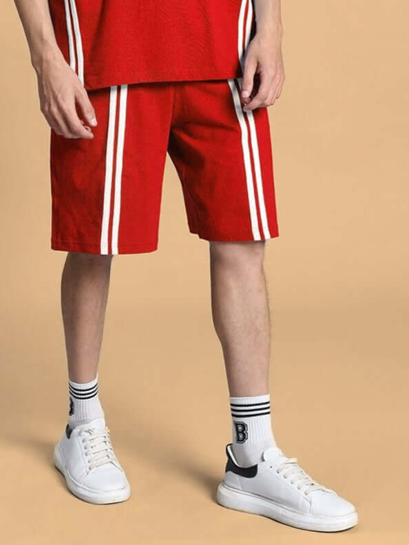 Man wearing Outlook regular fit red shorts with white stripes, styled with white sneakers and socks, standing against a beige background.
